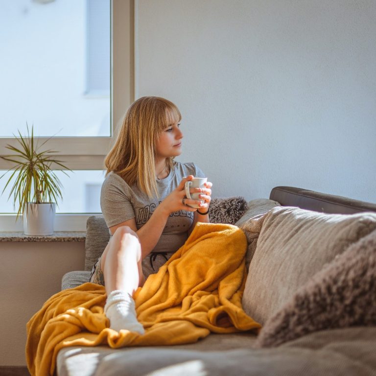 Wärme für Ihr Zuhause Eine Frau sitzt auf dem Sofa, hält eine Tasse und hat eine Decke über den Beinen.